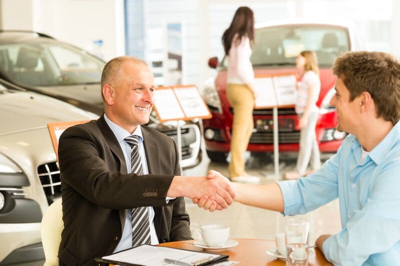 This photo shows a smiling man in a business suit shaking hands with a smiling man in a blue button down shirt as they sit at a table near some cars inside a dealership where a woman and a young girl are looking at a red car.