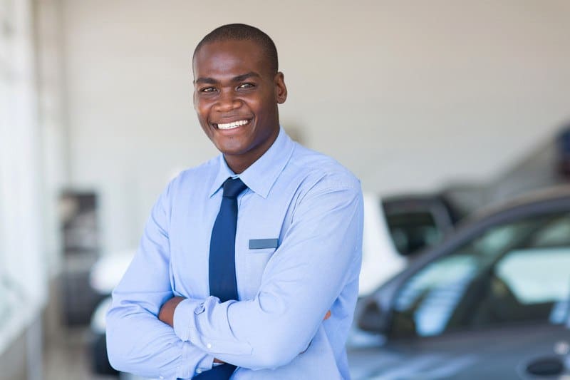 This photo shows a smiling man in a blue shirt and tie standing next to a gray car inside a car dealership.