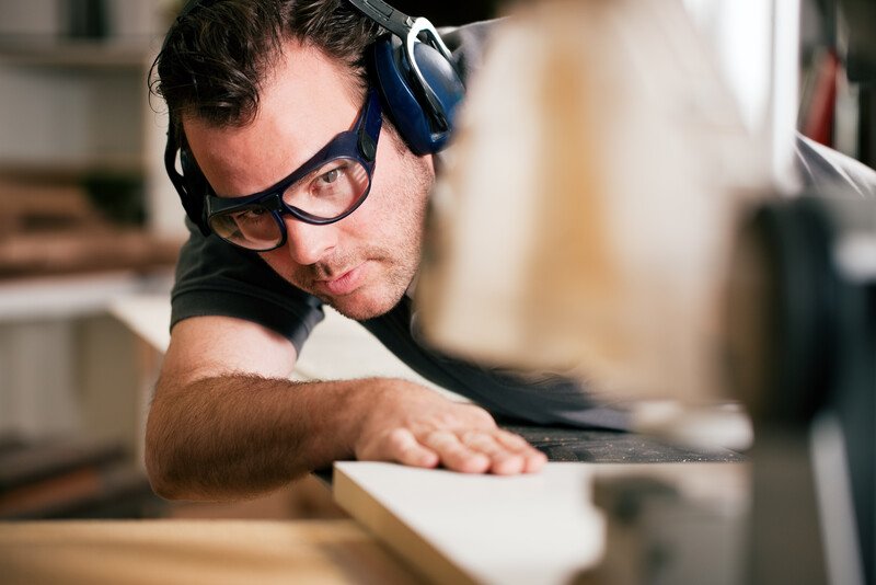 This photo shows a man in safety goggles lining up a board along a power saw.