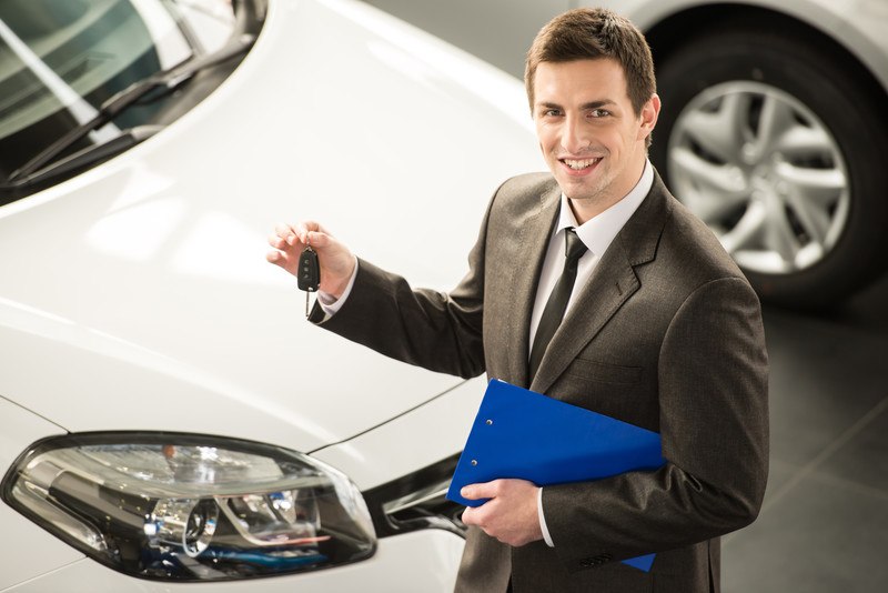 This photo shows a smiling young car salesman in a dark suit holding a set of keys as he stands next to a white car.