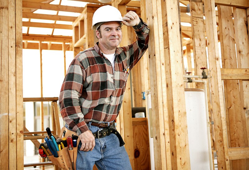 This photo shows a smiling man in a white hard hat and an orange and black plaid shirt with a tool belt leaning up against a framed wall in a new building, representing the question, do carpenters make good money?