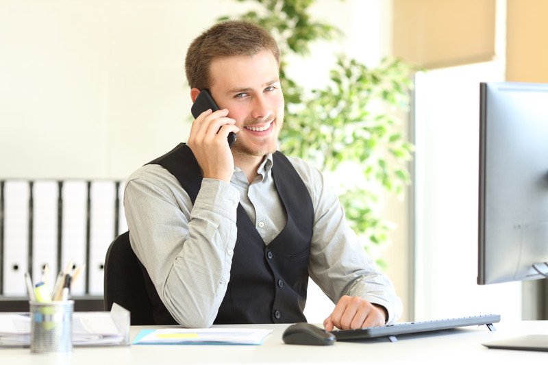 This photo shows a smiling young man dressed in a button down shirt and dress talking on the telephone with an open file in a brightly lit office, representing the question, do insurance adjusters make good money?