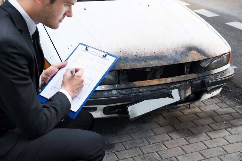 This photo shows a young man in a dark business suit looking at damage tot eh bumper of a white car as he writes on a paper held on a clipboard, representing an insurance adjuster at work.