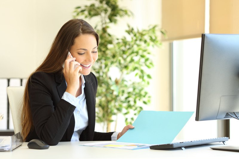 This photo shows a smiling young brunette woman in a business jacket looking at a file while talking on the telephone in a brightly lit office setting.