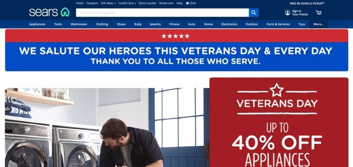 This screenshot of the home page for Sears has a dark blue header and search bar with white text above a red and blue announcement section, along with a photo of a man putting something into a washer in a laundry room on the left side of the page and a red and white sale announcement on the right side of the page.