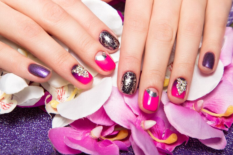 This photo shows a pair of female hands with purple, pink, and gold nail art on the fingernails, resting on a bed of pink silk flowers, representing the best nail art affiliate programs.