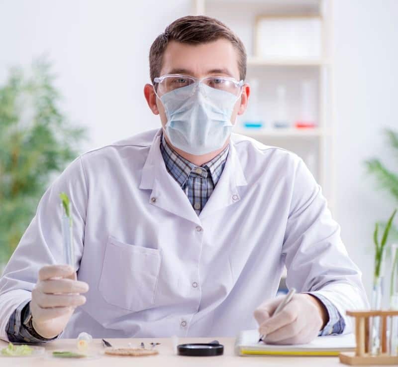 This photo shows a biochemist in a face mask and white lab coat, holding a test tube over a table with several scientific tools on it, while he writes something with his left hand.