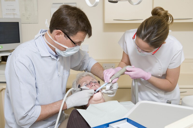 This photo shows a dentist in a blue and white striped shirt working in the mouth of a white haired elderly woman while a brunette dental assistant in a white shirt uses a suction hose in the woman's mouth.