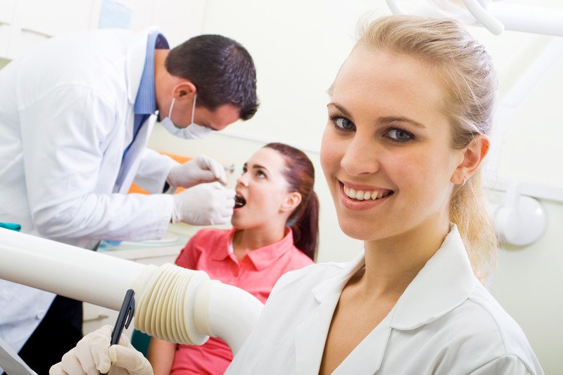 This photo shows a smiling blonde woman in a white lab coat, holding a clip board, while a dark haired dentist in a white lab coat and gloves looks into the mouth of a young woman patient.