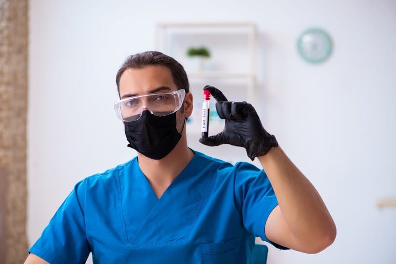 This photo shows a young man in safety glasses, a black face mask, black gloves, and blue scrubs, holding up a tube of blood in a clinical setting, representing the question, do biochemists make good money?