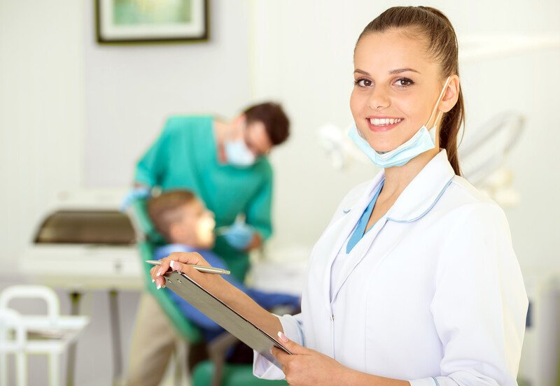 This photo shows smiling brunette woman in a white dental assistant jacket with a mask under her chin and holding a clipboard, while in the background a dentist in an aqua uniform shines a light into a patient's mouth, representing the question, do dental assistants make good money?