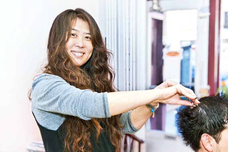 This photo shows a smiling brunette woman in a gray shirt and black apron cutting a customer's hair in a salon setting, representing the question, 'Do hair stylists make good money?'