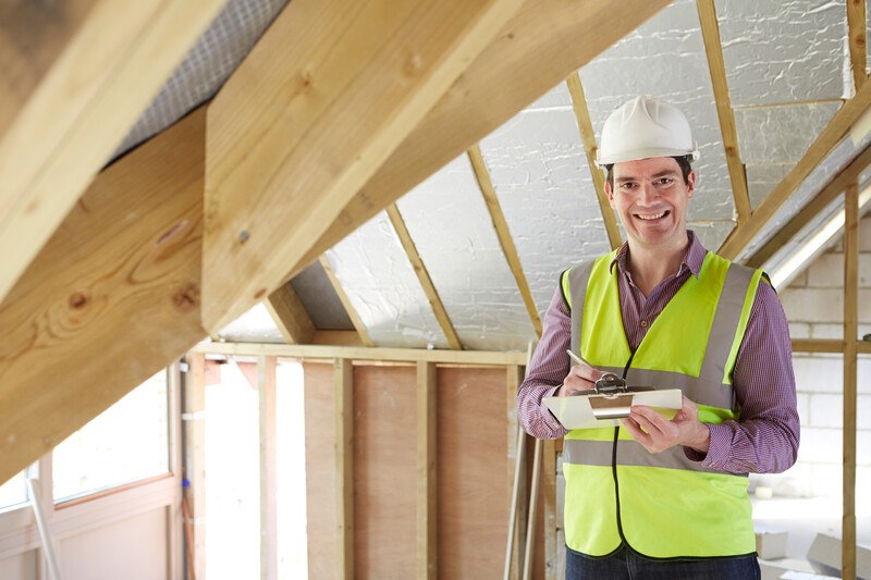 This photo shows a smiling man in a white hard hat, purple shirt, and yellow construction shirt writing on a clipboard near the attic ceiling of an unfinished home, representing the question, do home inspectors make good money?