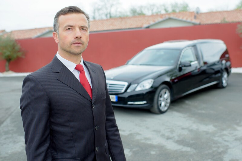 This photo shows a serious-faced man in a dark suit and red tie standing in front of a dark hearse in a parking lot, representing the question, do morticians make good money?