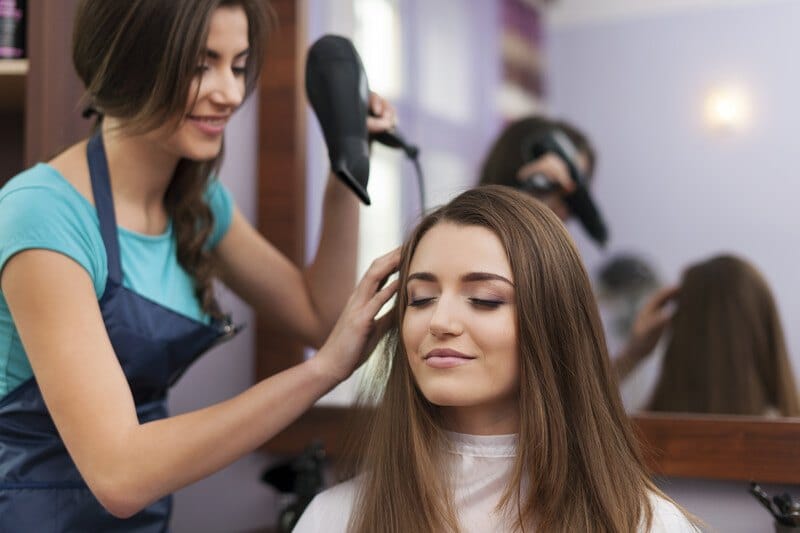 This photo shows a smiling dark haired woman in a blue apron and blue shirt blow drying the long brunette hair of another woman.