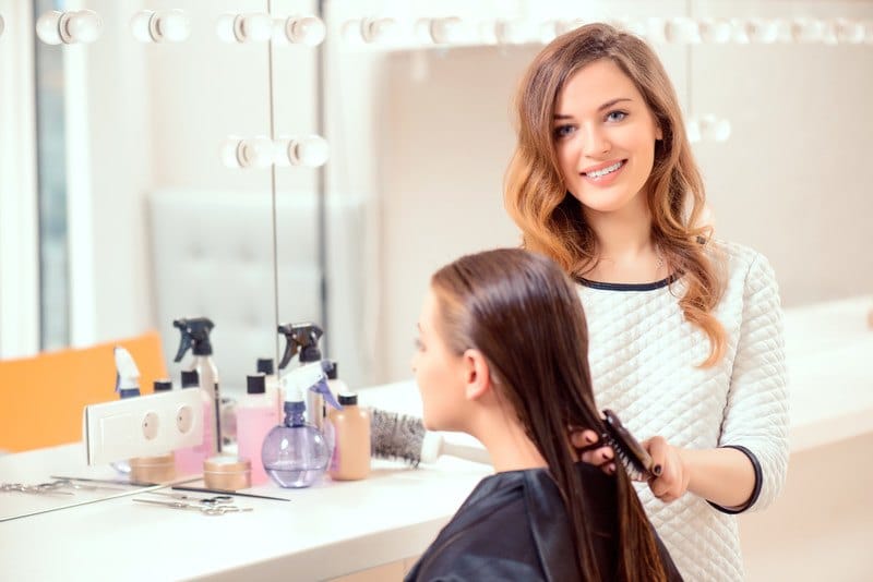 This photo shows a smiling hair stylist in a white shirt brushing through the long wet hair of a woman client in a black apron in a brightly lit salon setting.