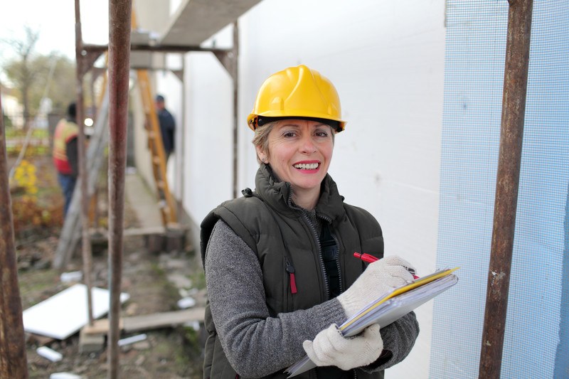 This photo shows a smiling woman in a yellow hard hat, a gray sweater and black coat vest, and white gloves, writing something on a yellow clipboard while construction workers work in the background.