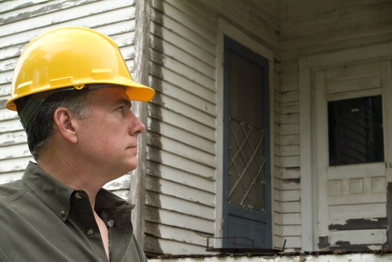 This photo shows a serious faced man in a yellow hard hat looking at the outside of a run-down white house.