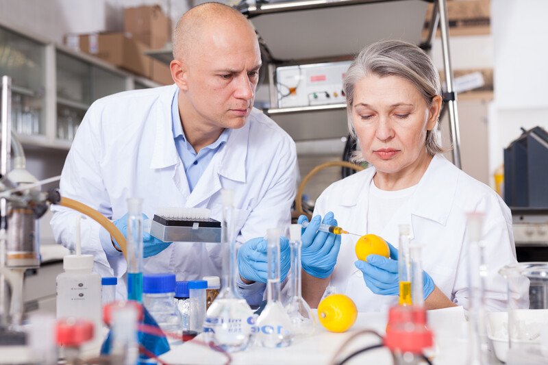 This photo shows a middle-aged man and a middle-aged woman, both in white lab coats and blue latex gloves, watching carefully as the woman injects something into a lemon in a laboratory setting.