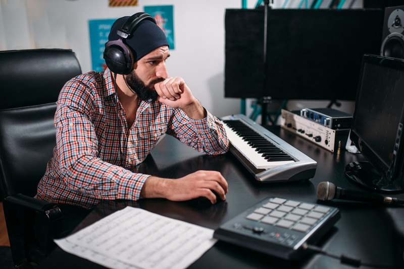 This photo shows a bearded man in a black beanie, headset, and multicolored button down shirt working on a computer near a piano keyboard, high tech equipment, and sheets of music.