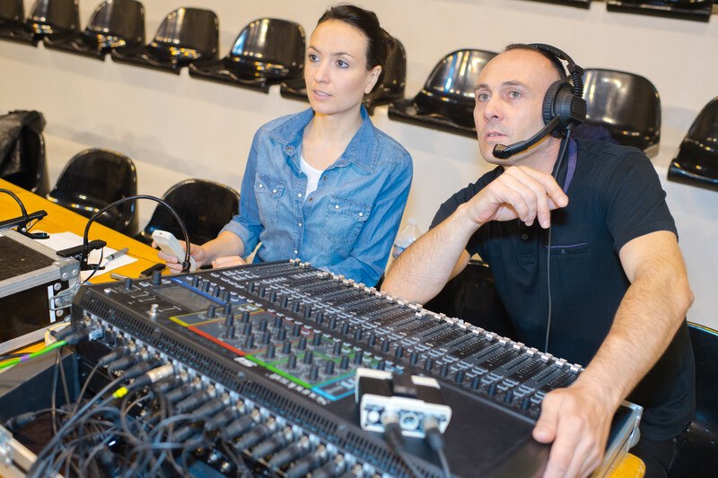 This photo shows a woman in a blue shirt and a man in a black shirt sitting in a recording studio behind high tech audio equipment.