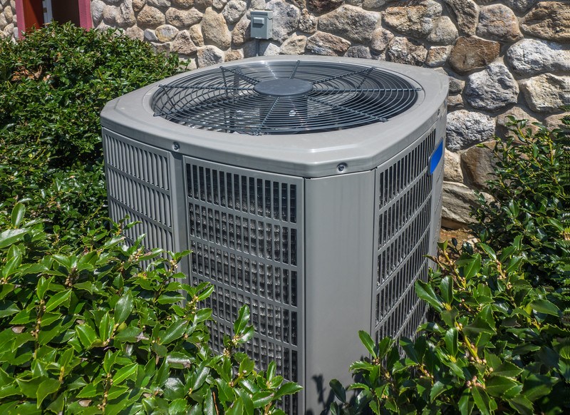 This photo shows a central air unit outside a rock-walled home, surrounded by Holly, representing the best HVAC affiliate programs.