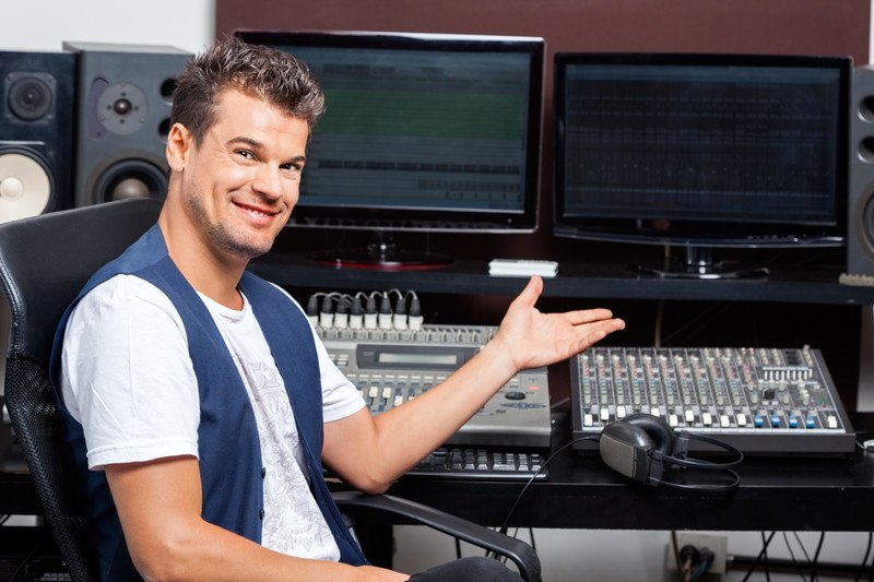 This photo shows a smiling man in a white tee shirt and blue vest holding a hand in front of high-tech audio equipment, representing the question, do audio engineers make good money?