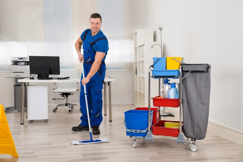 This photo shows a smiling man in a blue uniform, using a mop on a beige floor near a cleaning cart in an office building, representing the question, do janitors make good money?