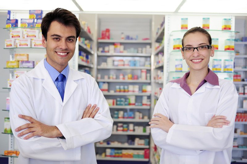 This photo shows a smiling man and a smiling woman in white lab coats, folding their arms and standing in front of shelves of medicines in a pharmacy, representing the question, do pharmacy techs make good money?