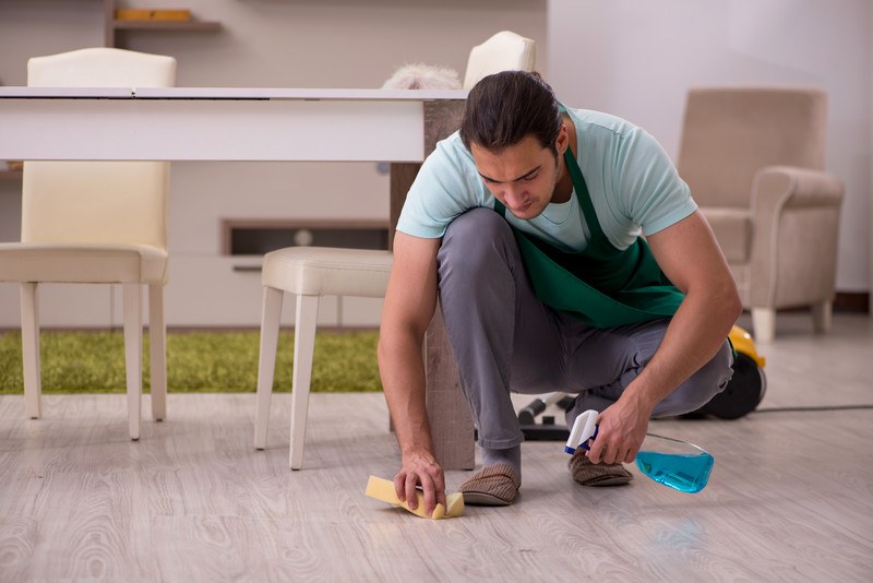 This photo shows a dark-haired man in a blue tee shirt, gray pants, and a green apron cleaning up a spill on a beige wooden floor with a spray bottle of window cleaner and a yellow sponge.