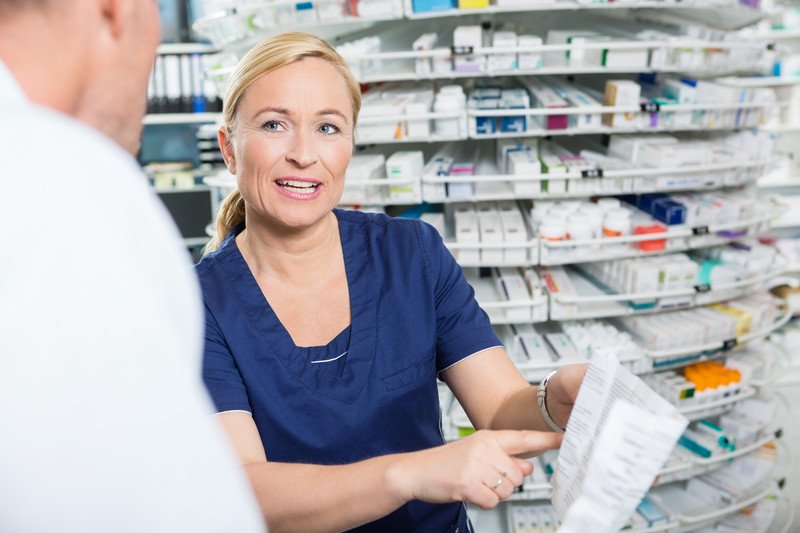 This photo shows a smiling blonde woman pharmacy tech talking to a pharmacist in a white coat about a prescription.