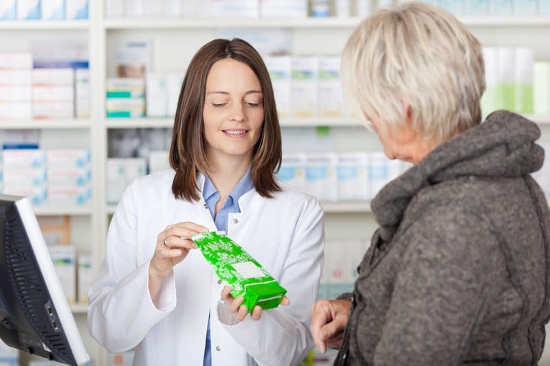 This photo shows a brunette woman pharmacy tech showing prescription in a green bag to a woman with short blonde hair and a brown coat.