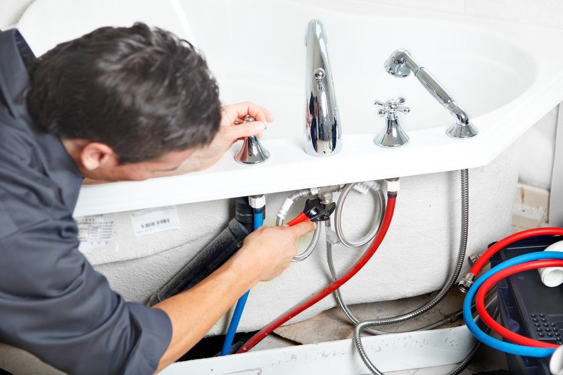 This photo shows the back view of a dark-haired plumber in charcoal-colored clothing using a wrench to tighten a pipe fitting on a bathtub that includes red and blue hoses under the lip of the tub.