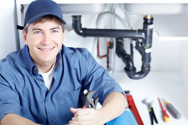 This photo shows a smiling man in a blue hat and blue clothes, holding a wrench and sitting in front of a set of tools and pipes under a sink.