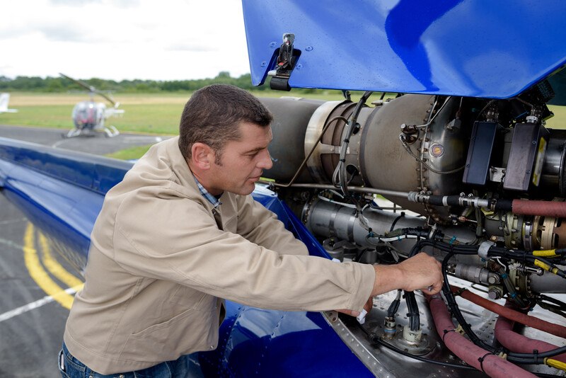 This photo shows a man in a tan jacket and jeans working on an open blue aircraft outdoors on an airfield.