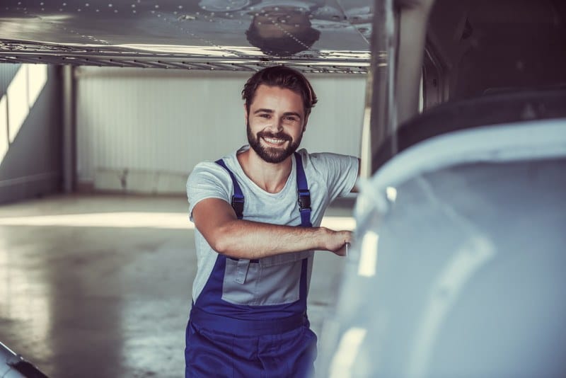 This photo shows a smiling bearded man in a gray tee shirt and blue coveralls standing beside a white aircraft in a hangar.