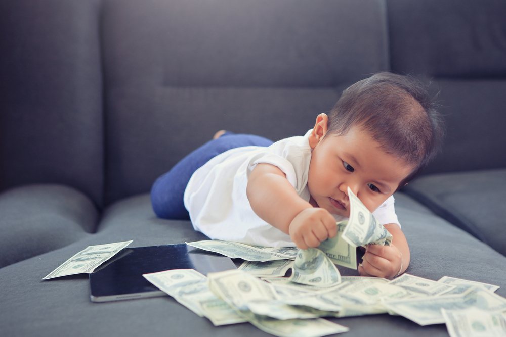 Asian toddler playing with US dollars on grey couch