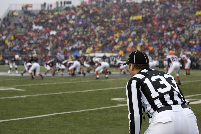 This photo shows a packed stadium, football players on the field, and a referee, representing the best college sports affiliate programs.