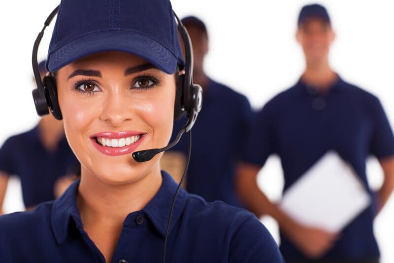 This photo shows a smiling dark-eyed woman in blue clothes, a blue hat, and a headset standing in front of a team of men in blue clothing.