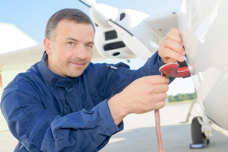 This photo shows a smiling man in a blue uniform working on an airplane using a red tool with a cord, representing the question, do aircraft mechanics make good money?