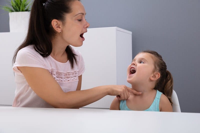 This photo shows a woman with a long dark ponytail, wearing a pink shirt, holding a finger on the throat of a smiling girl in a blue shirt, helping her feel her voice, as they sit at a white table, representing the question, do speech pathologists make good money?