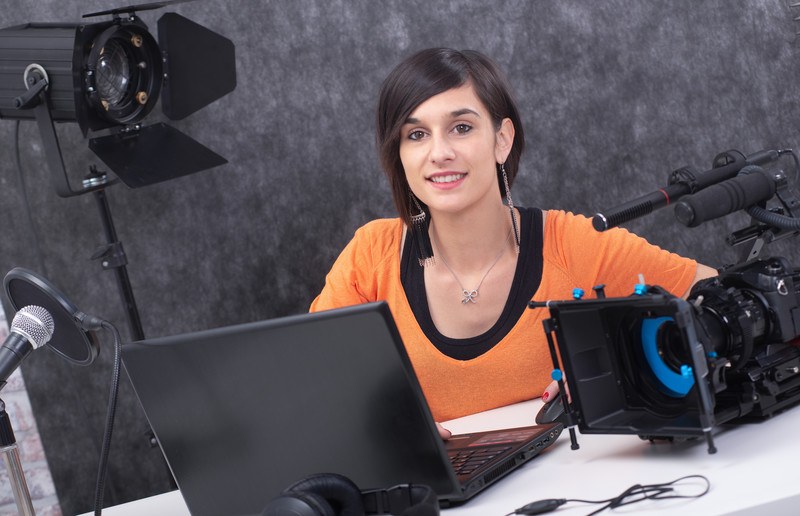 This photo shows a young woman with dark hair and an orange shirt sitting near a laptop and video equpiment, representing the question, do video editors make good money?