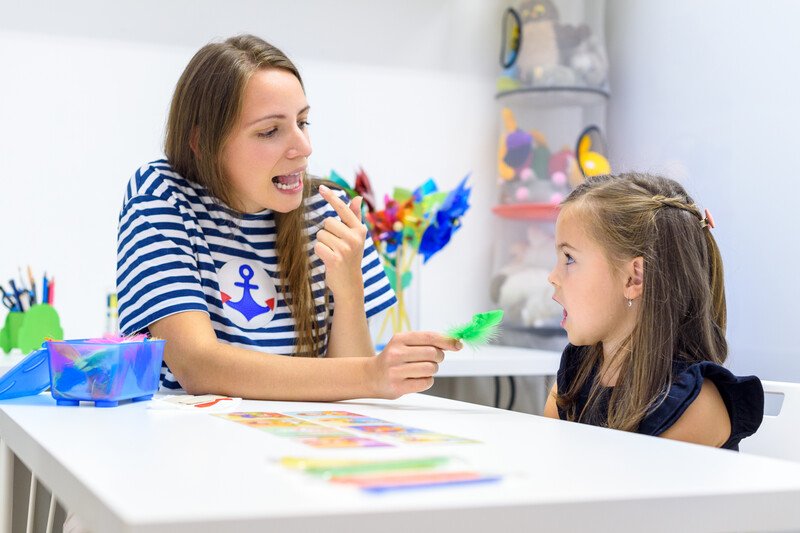 This photo shows a speech pathologist with long brown hair in a blue and white striped shirt showing a tongue position to a brunette girl in a white-walled classroom with multicolored toys and a white table.