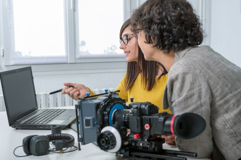 This photo shows a young woman with glasses and a yellow shirt with a curly-haired older woman in a gray suit jacket working with a pen tablet, laptop, and video equipment.