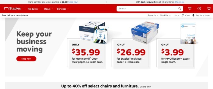 This screenshot of the home page for Staples has a gray header, a red navigation bar and search bar, a white announcement bar, and a gray main section with black text, product photos showing boxes of paper, and a red call to action button.