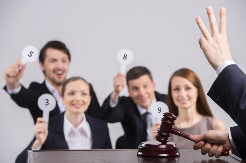 This photo shows four smiling men and women in business clothing holding paddles behind a table where an auctioneer's hand is showing three fingers, and his other hand is clapping a wooden mallet on its pedestal, representing the best auction affiliate programs.