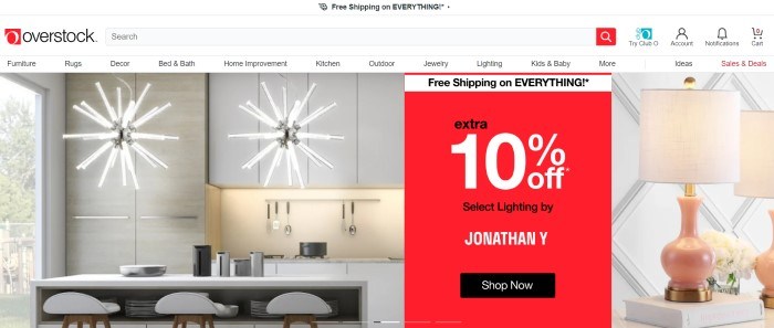 This screenshot of the home page for Overstock.com has a white header, navigation bar, and search bar above a photo on the left side of the page showing a kitchen with star-shaped chandeliers hanging above a white counter and white stools, and a photo of a lamp with a beige lampshade on the right side of the page, behind a red and white sales announcement.