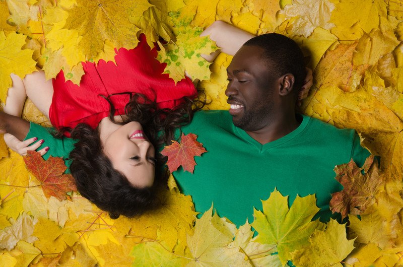 This photo shows a smiling dark skinned man in a green shirt and a smiling Asian woman in a red shirt playing together in a pile of yellow maple leaves, representing the best race dating affiliate programs.