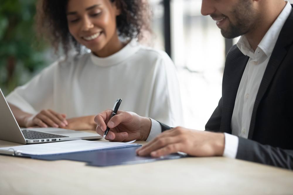 A man signs a document while a woman watches and smiles.