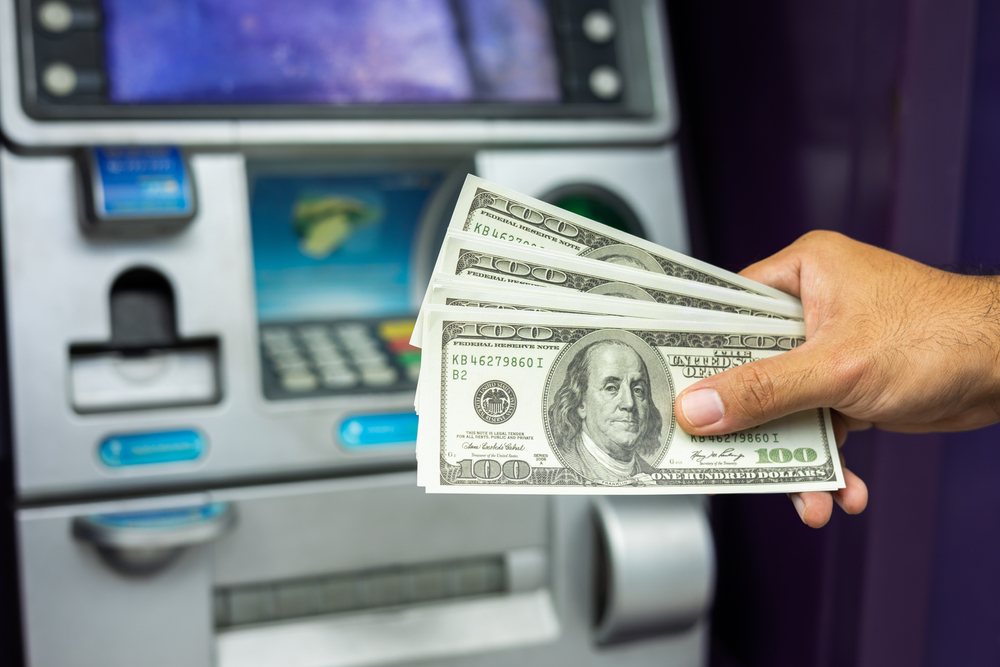 A man's hand holds a wad of hundred dollar bills in front of a freestanding ATM.